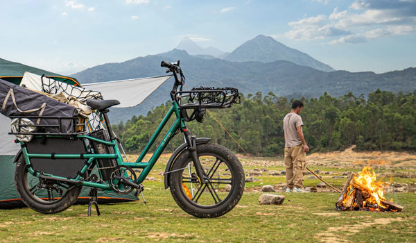 Fiido T2 E-Lastenrad mit Campingausrüstung beladen auf einem Bergcampingplatz neben einem Zelt und einem Lagerfeuer